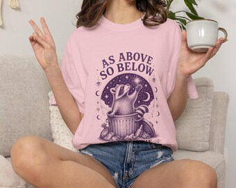 May include: A light pink t-shirt featuring a dark purple graphic of a raccoon sitting in a trash can, reaching toward a star. The text "AS ABOVE SO BELOW" is printed in an arch above the raccoon, surrounded by celestial symbols like moons and eyes.