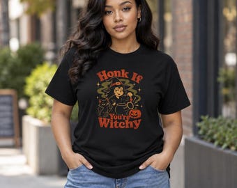 May include: A black short-sleeve t-shirt featuring a graphic print of a witch, a black cat, and a pumpkin. The text reads Honk If You're Witchy in red and orange lettering. The design includes small star accents and green smoke details.