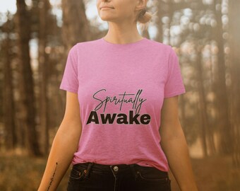 May include: A pink t-shirt with the words "Spiritually Awake" in black script and block letters. The shirt is worn with black jeans. The image is taken outdoors with a blurred forest background.