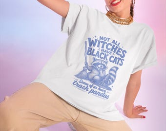 May include: A white short-sleeved t-shirt featuring a blue graphic of a raccoon wearing a witch hat and holding a broom. The text reads: NOT ALL WITCHES HAVE BLACK CATS, Some of us have trash pandas. The design includes small star accents.
