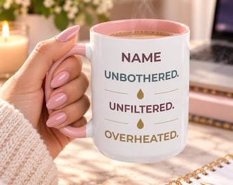 May include: A white ceramic mug with a soft pink interior and handle. The front features the text NAME UNBOTHERED. UNFILTERED. OVERHEATED. in dark blue, maroon, and gold, separated by small gold droplet icons and horizontal lines.
