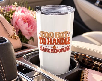 May include: A white stainless steel tumbler with a clear plastic lid featuring the orange and red text "TOO HOT TO HANDLE BLAME MENOPAUSE" with a small flame icon centered between the two lines of text on the front of the cup.