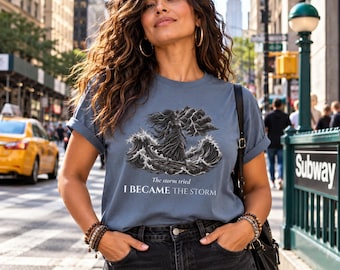 May include: A slate blue short-sleeve t-shirt featuring a graphic of a figure standing amidst turbulent ocean waves and storm clouds. The text printed below the illustration reads, The storm tried I BECAME THE STORM.