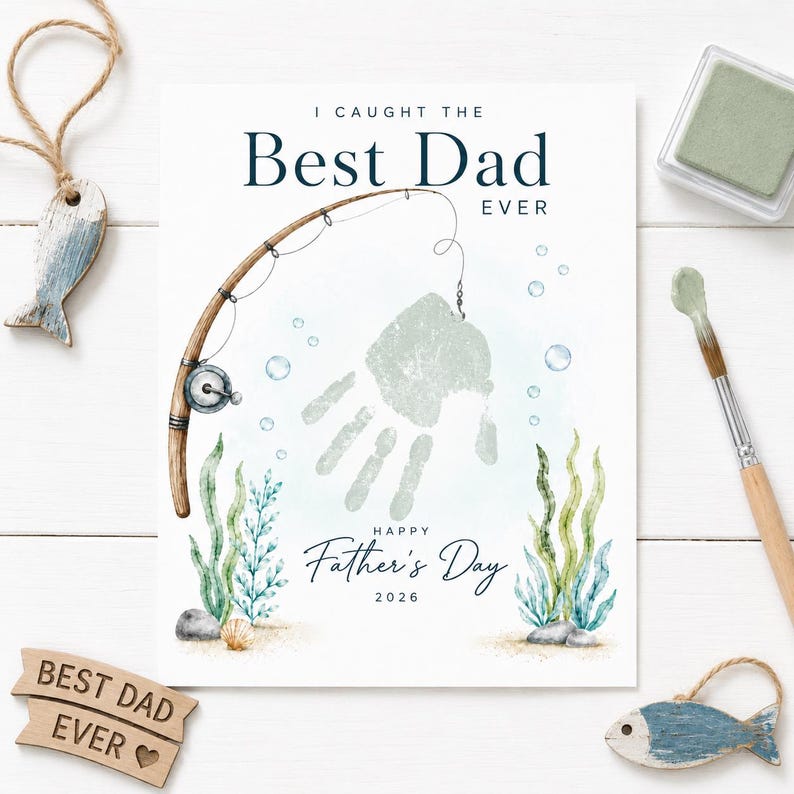 May include: A Father's Day print with the text "I Caught the Best Dad Ever" and "Happy Father's Day 2026". The design includes a fishing rod, a handprint, and aquatic plants. Decorative wooden fish and a stamp pad are also visible.
