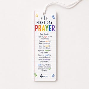 Op de afbeelding: Een witte rechthoekige bladwijzer met een lijntjespatroon, voorzien van de tekst FIRST DAY PRAYER in regenboogkleuren. Het bevat een kruisicoon, kleurrijke sterren en paperclips, met een gebed in zwarte tekst en een wit koord aan de bovenkant.