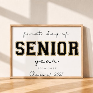 May include: A rectangular wooden frame containing a white sign with black and gold text. The text reads first day of SENIOR year 2026-2027 Class of 2027. The word SENIOR is in a large, bold, varsity-style font with black fill and gold outlines.