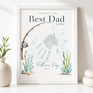 May include: A framed print with the text "I Caught The Best Dad Ever" and "Happy Father's Day 2026". The artwork features a fishing rod, a handprint, bubbles, and aquatic plants in shades of blue, green, and brown. The frame is white.
