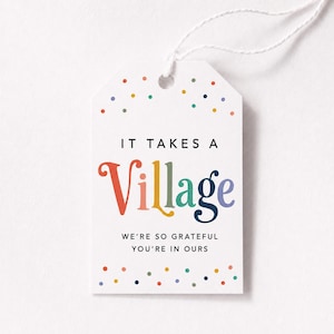 Puede incluir: Etiqueta de regalo blanca con el texto "IT TAKES A Village" en negro y letras coloridas. La palabra "Village" está en colores del arcoíris. La etiqueta también incluye el texto "WE'RE SO GRATEFUL YOU'RE IN OURS". La etiqueta está decorada con lunares de colores y tiene una cuerda blanca.