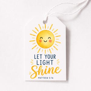 Op de afbeelding: Een wit rechthoekig cadeaulabel met een aquarelillustratie van een lachende gele zon met oranje wangen. De tekst luidt LET YOUR LIGHT SHINE in donkerblauw en geel, gevolgd door de kleine zwarte tekst MATTHEW 5:16 aan de onderkant.