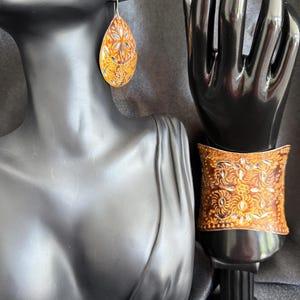 May include: A pair of teardrop-shaped earrings and a cuff bracelet. The jewelry is a warm orange color with intricate floral patterns in gold and blue. The earrings hang from silver hooks. The cuff bracelet is displayed on a black hand.