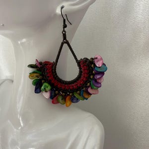 May include: Handmade, bohemian-style earrings. The earrings feature a dark brown macrame design with a red center and are adorned with colorful, iridescent shell accents. The earrings hang from dark metal hooks.