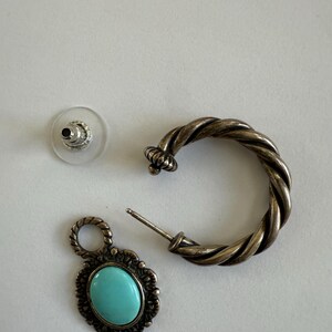 May include: A silver-toned earring set featuring a twisted hoop earring, a turquoise-colored oval pendant earring, and a clear earring back. The pendant earring has a decorative border.
