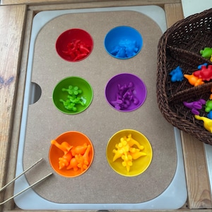 May include: A set of six colourful bowls filled with small plastic dinosaurs. The bowls are arranged in a row on a wooden surface. A pair of tongs is lying on the surface. A wicker basket filled with more dinosaurs is in the top right corner of the image.