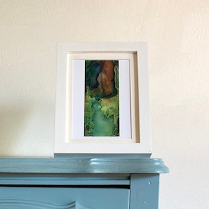 Puede incluir: Una pintura abstracta enmarcada con colores verde, azul y marrón. La pintura está en un marco blanco y está colgada en una pared azul claro.