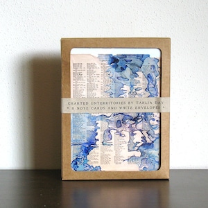 Peut inclure: Un ensemble de 8 cartes de vœux avec un design abstrait aquarelle bleu et blanc sur une page de dictionnaire vintage. Les cartes sont emballées dans une enveloppe brune avec le texte "CHARTERED UNTERRITORIES BY TAHLIA DAY * 8 NOTE CARDS AND WHITE ENVELOPES *"