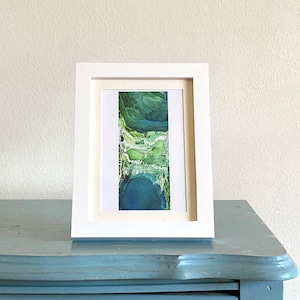 Puede incluir: Una pintura abstracta enmarcada con tonos de verde y azul. La pintura está en un marco blanco y está sobre una superficie azul.