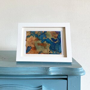 Può includere: Un dipinto astratto ad acquerello incorniciato con tonalità di blu, marrone e verde. Il dipinto è in una cornice bianca e si trova su una superficie blu.