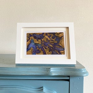 Peut inclure: Une impression d'art abstrait encadrée avec un schéma de couleurs or et violet. L'impression est dans un cadre blanc et repose sur une surface bleue.