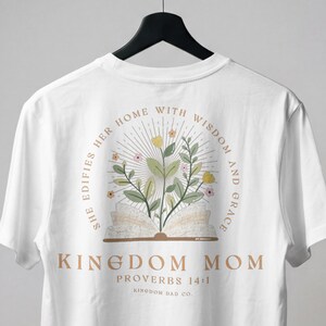 Op de afbeelding: Wit T-shirt met een bloemenontwerp en de tekst "KINGDOM MOM". Het ontwerp bevat een open boek met bloemen en de woorden "SHE EDIFIES HER HOME WITH WISDOM AND GRACE" in een schreefletter.