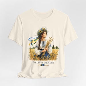 Peut inclure: Un t-shirt à manches courtes de couleur crème avec une illustration graphique représentant une personne portant une couronne de fleurs et des vêtements traditionnels, tenant du blé dans un champ. Le texte "FIELD OF MEMORY" est imprimé en dessous.