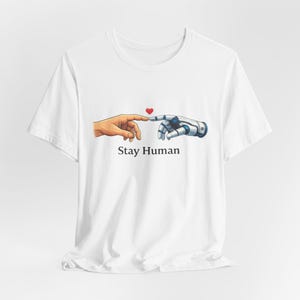 Könnte beinhalten: Weißes T-Shirt mit einer Grafik, die eine menschliche Hand und eine Roboterhand zeigt, die sich fast berühren, mit einem roten Herzen darüber. Der Text "Stay Human" ist darunter aufgedruckt. Das T-Shirt besteht aus weißem Stoff.