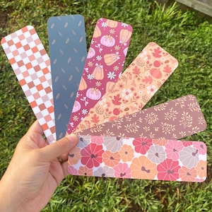 Puede incluir: Una colecci&oacute;n de seis marcap&aacute;ginas decorativos con varios dise&ntilde;os tem&aacute;ticos de oto&ntilde;o. Los dise&ntilde;os incluyen calabazas, hojas y arreglos florales en tonos naranja, rosa y marr&oacute;n. Un marcap&aacute;ginas presenta un patr&oacute;n de cuadros.