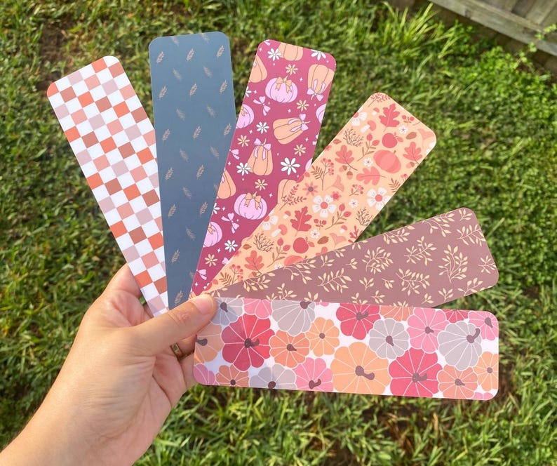 Puede incluir: Una colecci&oacute;n de seis marcap&aacute;ginas decorativos con varios dise&ntilde;os tem&aacute;ticos de oto&ntilde;o. Los dise&ntilde;os incluyen calabazas, hojas y arreglos florales en tonos naranja, rosa y marr&oacute;n. Un marcap&aacute;ginas presenta un patr&oacute;n de cuadros.