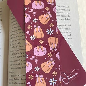 Puede incluir: Marcap&aacute;ginas burdeos con un patr&oacute;n repetido de calabazas naranjas y rosas, lazos y flores blancas. El texto "Pumpkin Bows" est&aacute; en la parte superior. La parte posterior del marcap&aacute;ginas tiene el logotipo de Omnia Designs.