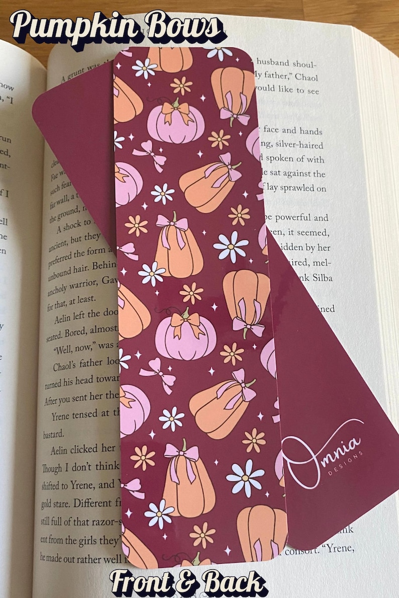 Puede incluir: Marcap&aacute;ginas burdeos con un patr&oacute;n repetido de calabazas naranjas y rosas, lazos y flores blancas. El texto "Pumpkin Bows" est&aacute; en la parte superior. La parte posterior del marcap&aacute;ginas tiene el logotipo de Omnia Designs.