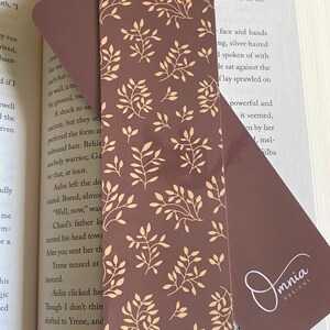 Puede incluir: Un marcap&aacute;ginas marr&oacute;n con un dise&ntilde;o floral beige. El marcap&aacute;ginas es rectangular con esquinas redondeadas y el texto "Front & Back". El logotipo "Omnia Designs" es visible.