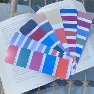 May include: An open book with pages displaying text, accompanied by a fan of color swatches. The swatches showcase a variety of colors, including shades of blue, pink, and orange. The book is open on a woven chair.