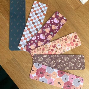 Puede incluir: Un conjunto de marcap&aacute;ginas rectangulares con varios dise&ntilde;os. Los dise&ntilde;os incluyen un verde oscuro con detalles de hojas, un patr&oacute;n de cuadros, calabazas, arreglos florales y un patr&oacute;n floral en tonos rosas, naranjas y marrones. Dispuestos sobre una superficie de madera.