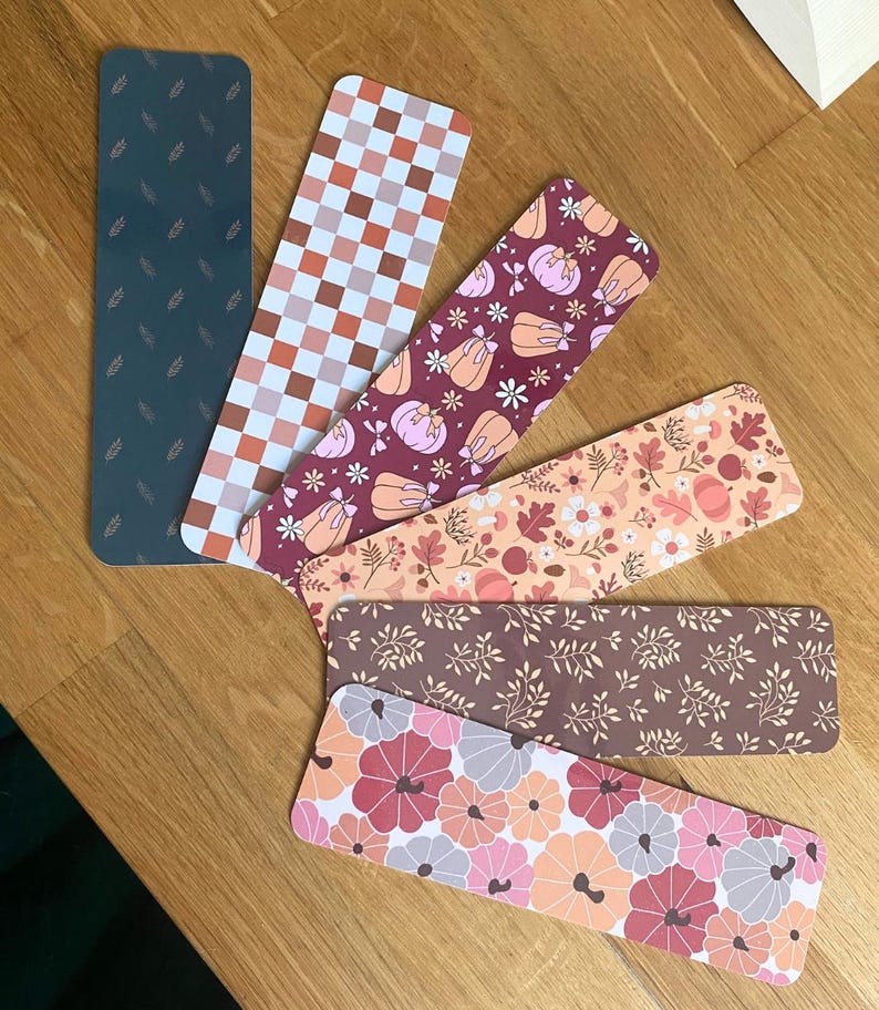 Puede incluir: Un conjunto de marcap&aacute;ginas rectangulares con varios dise&ntilde;os. Los dise&ntilde;os incluyen un verde oscuro con detalles de hojas, un patr&oacute;n de cuadros, calabazas, arreglos florales y un patr&oacute;n floral en tonos rosas, naranjas y marrones. Dispuestos sobre una superficie de madera.