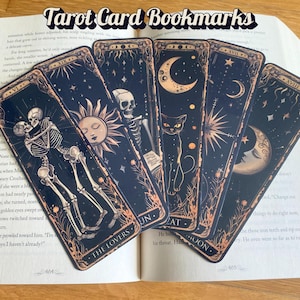 May include: A collection of tarot card bookmarks with celestial and gothic illustrations. Each bookmark features a different card design, including The Lovers, Sun, Cat, and Moon. The bookmarks are displayed on an open book with the text "Tarot Card Bookmarks" at the top.
