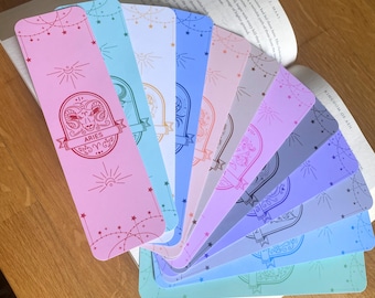 May include: A collection of colorful, illustrated bookmarks fanned out on a wooden surface. Each bookmark features a different pastel color and a zodiac sign design, including the Aries symbol. The bookmarks are decorated with stars and sunbursts.
