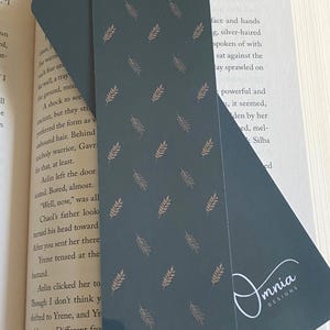 Puede incluir: Dos marcap&aacute;ginas de color verde azulado oscuro con un patr&oacute;n repetido de ramitas doradas. El texto "Evergreen Sprigs" est&aacute; en la parte superior y "Front & Back" en la inferior. Un marcap&aacute;ginas est&aacute; insertado en un libro.