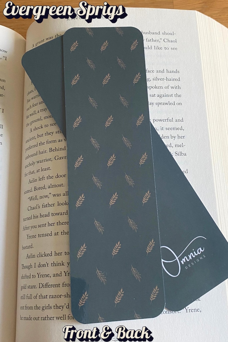 Puede incluir: Dos marcap&aacute;ginas de color verde azulado oscuro con un patr&oacute;n repetido de ramitas doradas. El texto "Evergreen Sprigs" est&aacute; en la parte superior y "Front & Back" en la inferior. Un marcap&aacute;ginas est&aacute; insertado en un libro.