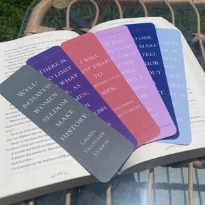 May include: An open book with several colorful bookmarks. The bookmarks are in shades of gray, purple, red, lavender, and blue, each with a quote. The gray bookmark reads "Well-behaved women seldom make history."