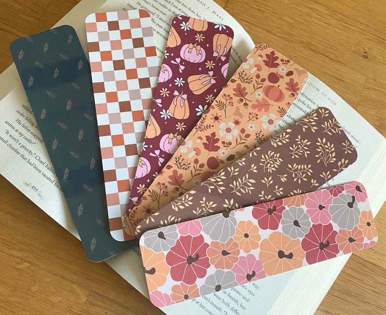 Puede incluir: Colecci&oacute;n de seis marcap&aacute;ginas decorativos con dise&ntilde;os oto&ntilde;ales. Los dise&ntilde;os incluyen detalles de hojas, un patr&oacute;n de cuadros, calabazas y motivos florales en tonos naranja, marr&oacute;n y rosa. Los marcap&aacute;ginas est&aacute;n colocados sobre un libro abierto.