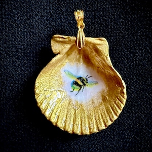 May include: A gold-painted seashell pendant with a painted bee design. The pendant has a gold bail for hanging. The bee is black and yellow with blue and green accents, set against a white background.