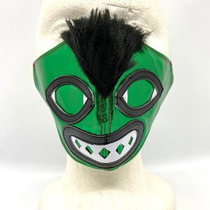 May include: Green wrestling mask with black and white details. The mask has a black fur tuft on top and a wide, toothy grin.
