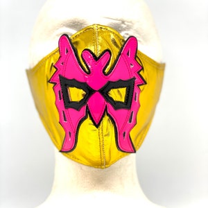 May include: A gold wrestling mask with a pink butterfly design. The mask has black accents around the eyes and a black outline around the butterfly.