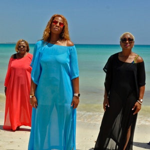 May include: Six women wearing colorful beach cover-ups stand on a sandy beach with the ocean in the background. The cover-ups are in shades of blue, white, black, and red. The women are wearing sunglasses and watches.
