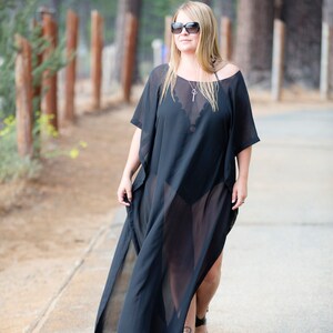 May include: A black, sheer, flowing cover-up dress with a split front, worn over a black swimsuit. The dress has short sleeves and is paired with black sandals. The person is wearing sunglasses and a necklace.
