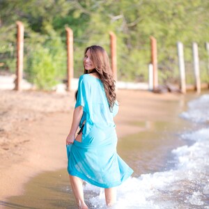 May include: A light blue beach cover-up with a flowing design. The cover-up is being held up as the person walks through the water. The background includes a sandy beach, water, and a green treeline.