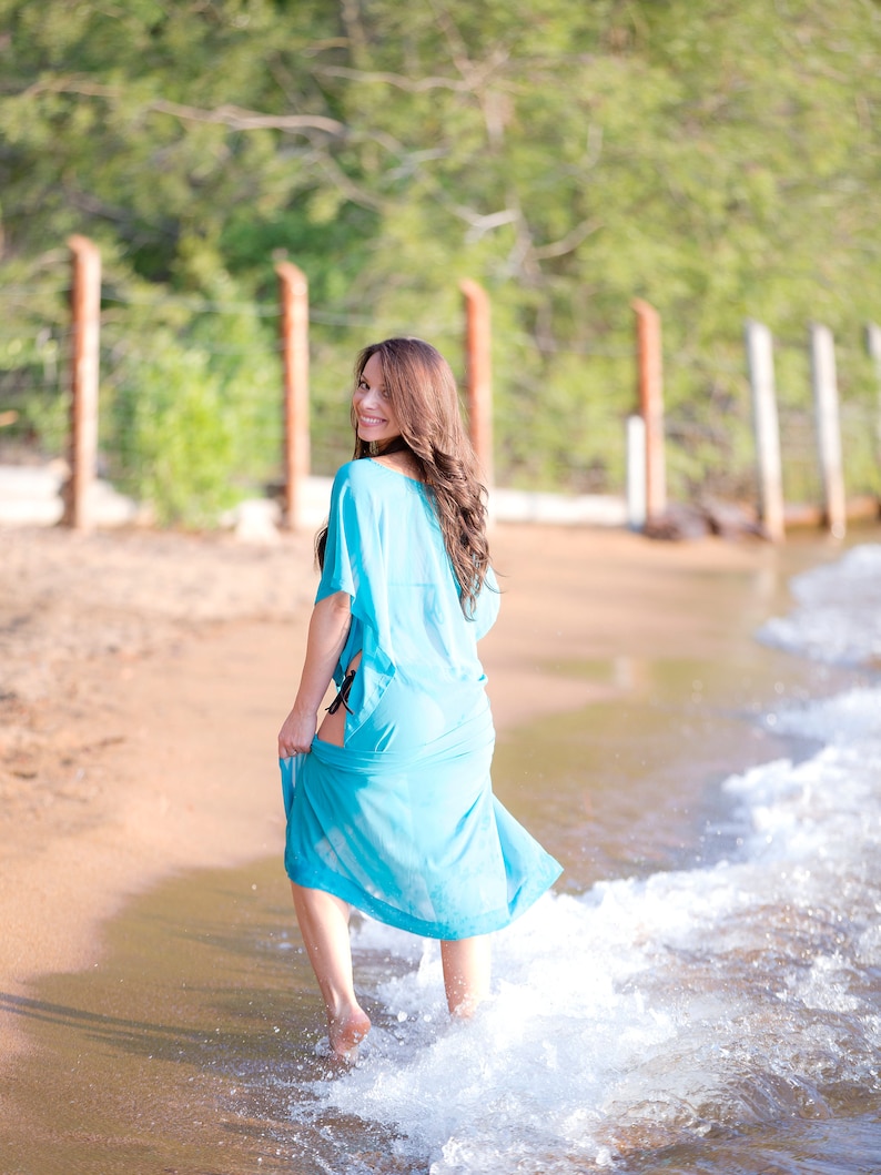 May include: A light blue beach cover-up with a flowing design. The cover-up is being held up as the person walks through the water. The background includes a sandy beach, water, and a green treeline.