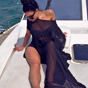 May include: A woman wearing a black off-the-shoulder cover-up and sunglasses sits on a white boat deck. The sheer fabric drapes over her, revealing a black bikini. The background shows a cityscape and the ocean.