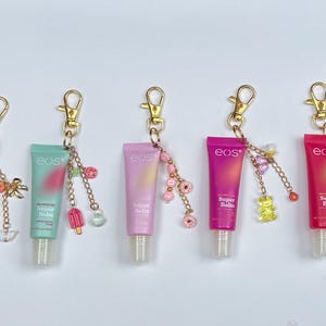 May include: Five eos Super Balm lip balm keychains in pink, teal, and purple tubes with gold-colored clasps and decorative charms. Charms include bows, ice pops, donuts, and gummy bears.