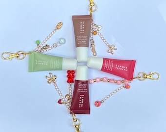May include: Four Summer Fridays lip butter balm tubes in beige, green, pink, and dark red, each attached to a gold-tone metal keychain. The keychains feature various charms including a red gummy bear, a bow, a seahorse, and beads spelling HANNAH.