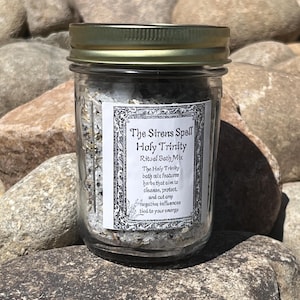 May include: A clear glass jar with a gold lid, filled with a mix of herbs and salts. The jar has a label that reads "The Sirens Spell Holy Trinity Ritual Bath Mix." The jar is sitting on a bed of rocks.